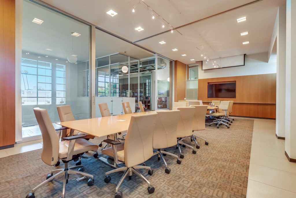 Barrister Suites Office Lease Modern conference room with a long wooden table, beige office chairs on wheels, glass walls, carpeted floor, and bright overhead lighting. Large windows let in natural light, and there is a TV mounted on a wooden wall.