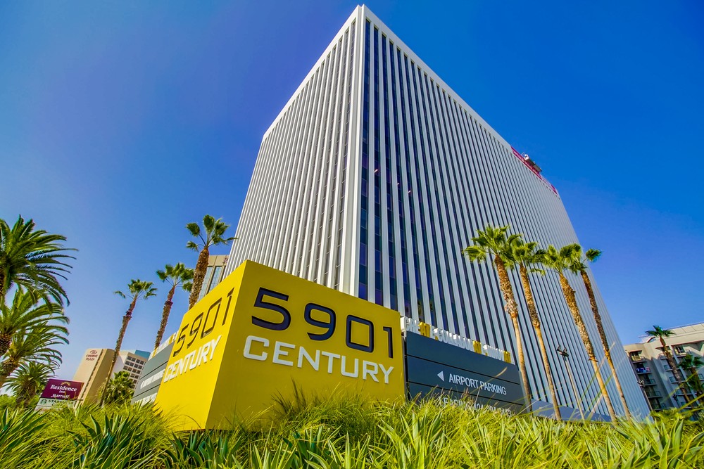 Barrister Suites Office Lease Tall modern office building with vertical lines, surrounded by palm trees and greenery. A large yellow sign in front reads 5901 Century. The sky is clear and blue.