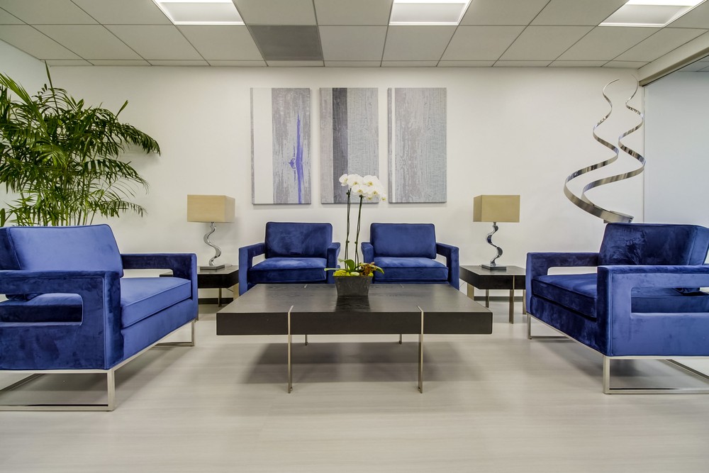 Barrister Suites Office Lease Modern office waiting area with four blue velvet armchairs around a dark coffee table, two table lamps, a tall plant, white orchid centerpiece, abstract wall art, and a metal sculpture in the background.