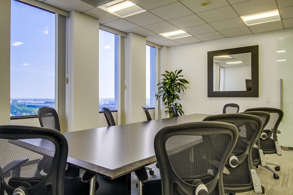 Barrister Suites Office Lease A modern conference room with a long table, several black office chairs, large windows letting in natural light, a potted plant, and a wall mirror.