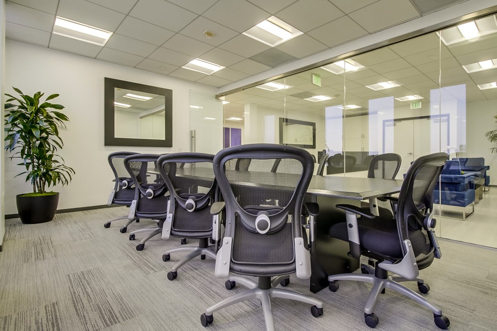 Barrister Suites Office Lease Modern conference room with a long table, eight black mesh office chairs, a large wall mirror, potted plant, glass wall, and bright overhead lighting. The space is clean and minimalist.