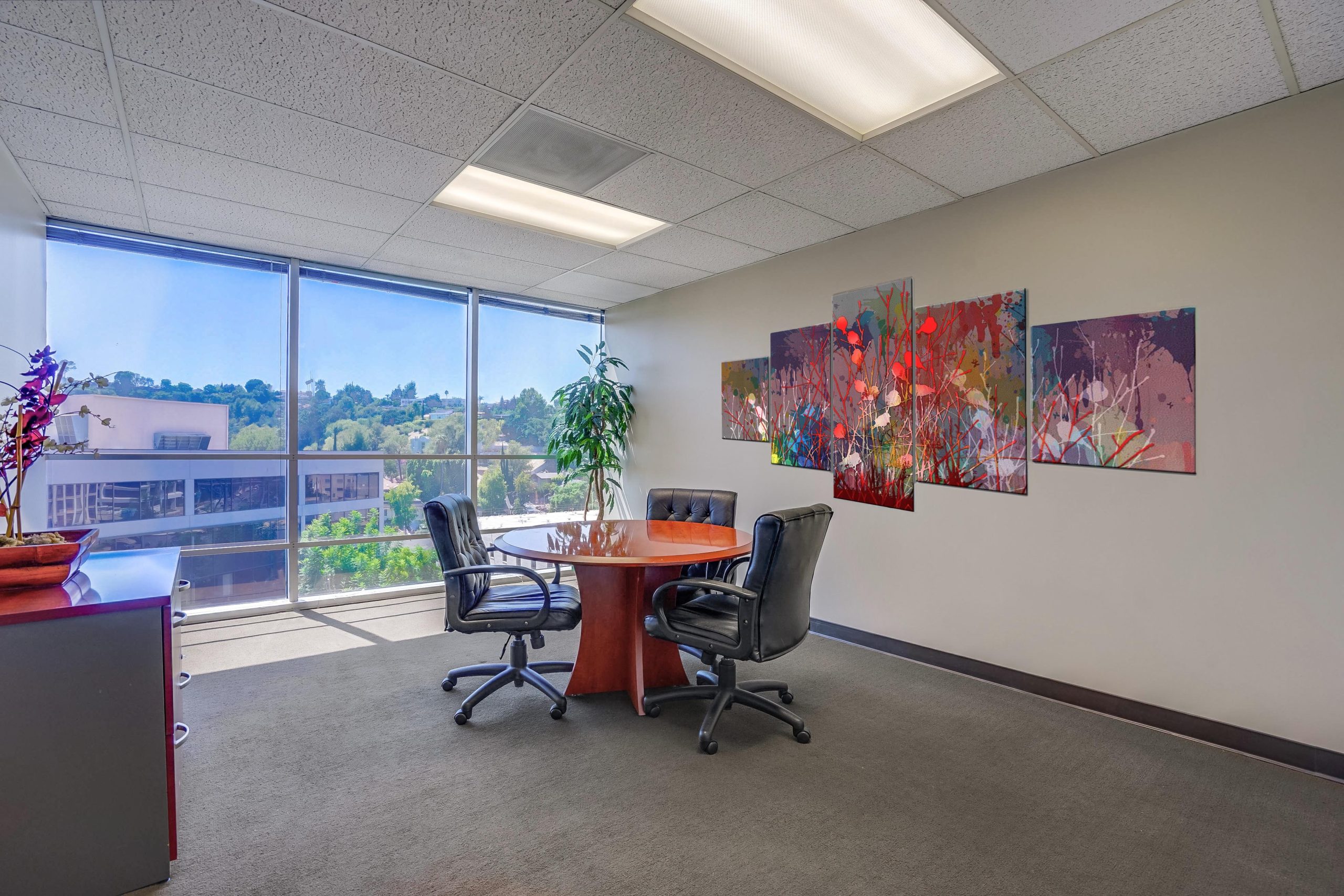 Barrister Suites Office Lease A small, modern office meeting room with a round wooden table, four black chairs, abstract wall art, a potted plant, and large windows overlooking greenery and buildings.