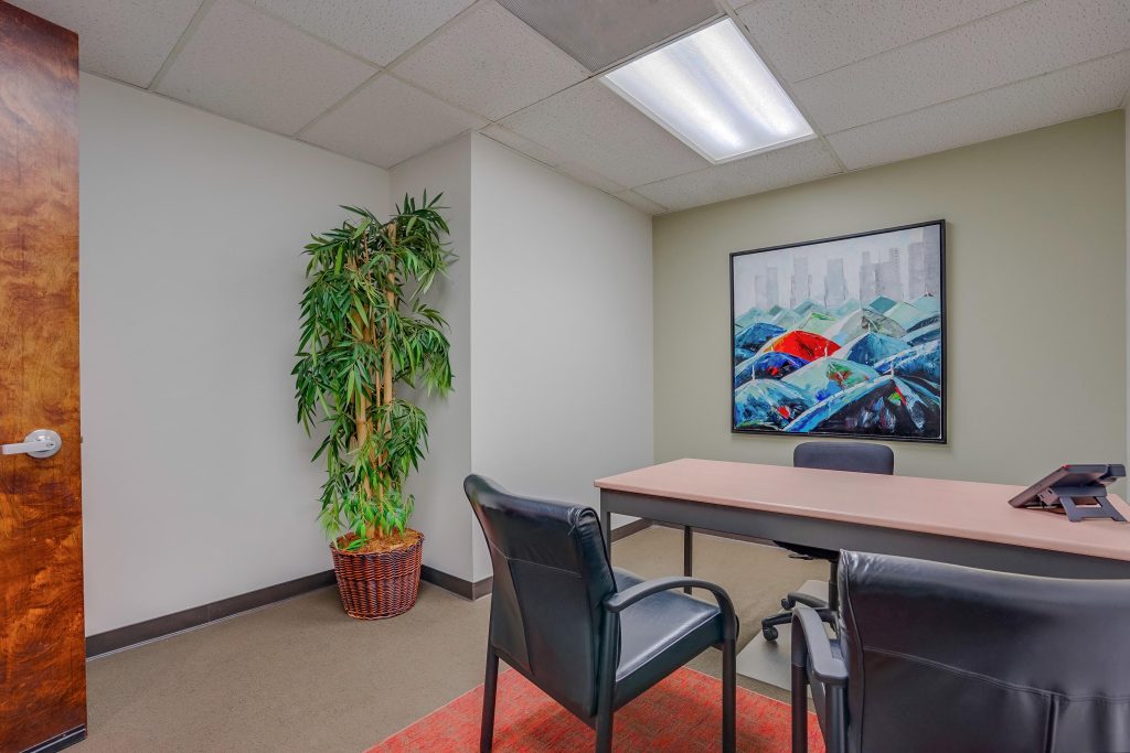 Barrister Suites Office Lease A small office with two black chairs, a desk with a phone, a potted plant in the corner, a modern painting on the wall, and bright overhead lighting.