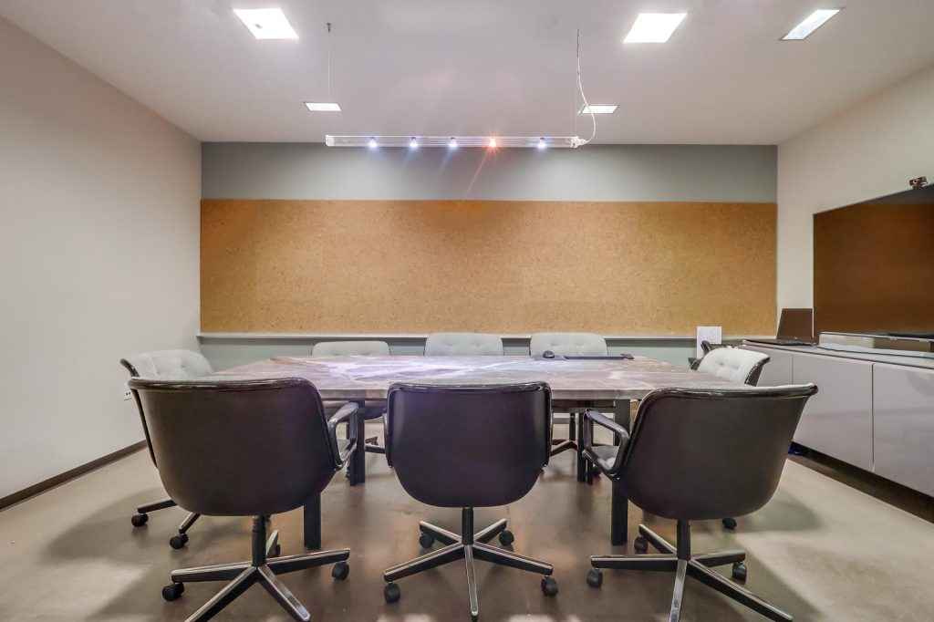 Barrister Suites Office Lease A modern conference room with eight gray chairs around a rectangular table, a large corkboard on the wall, overhead lighting, and cabinets in the corner. The room has a neutral color scheme.