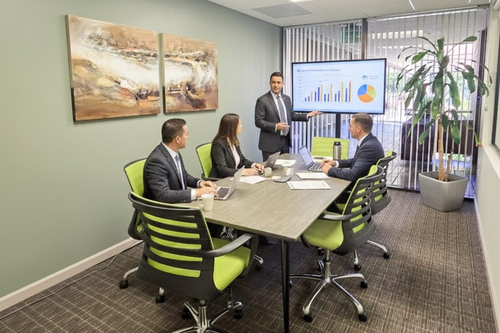 Barrister Suites Office Lease Four people in business attire sit around a conference table in a modern office as one person stands, presenting a bar and pie chart on a screen. Laptops and papers are on the table, and a plant is in the corner.