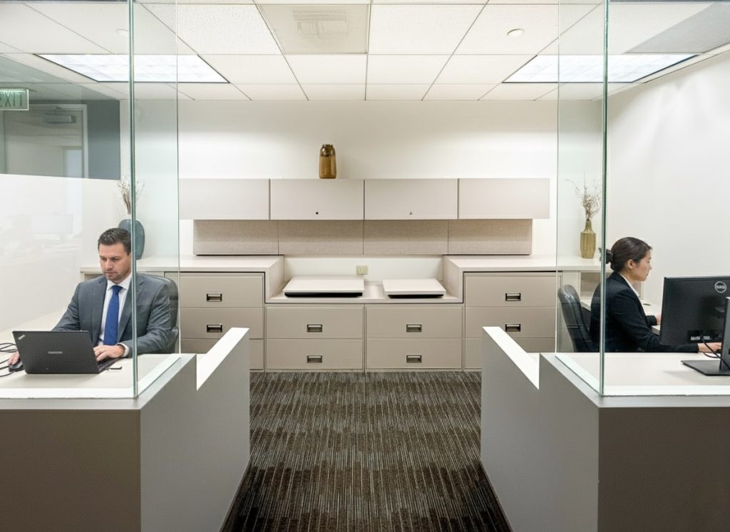 Barrister Suites Office Lease Two people work at separate desks in a modern office, divided by glass partitions. Both use computers, and neutral-colored cabinets and decor are visible in the background. The office is well-lit and orderly.
