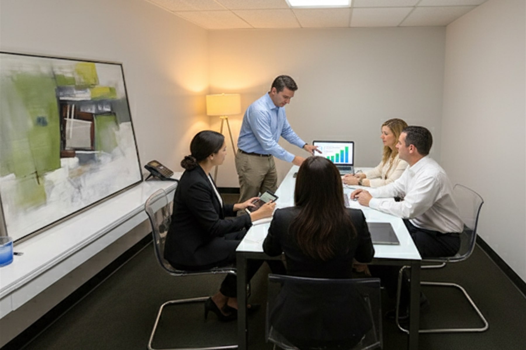 Barrister Suites Office Lease Four people sit at a conference table while one man stands and points at a computer screen displaying a bar chart. The room is modern with abstract art on the wall and a lamp in the corner.