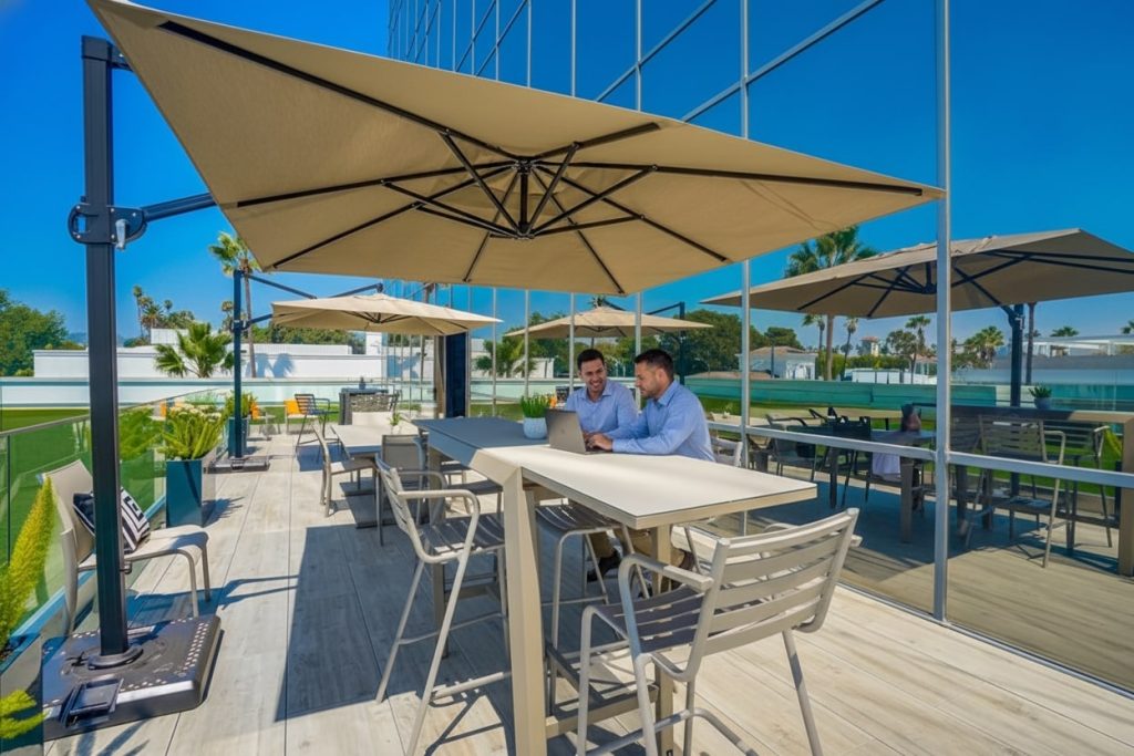 Barrister Suites Office Lease Two men sit and talk at a tall outdoor table on a modern patio with large umbrellas, surrounded by empty chairs and greenery, next to a building with glass walls on a sunny day.