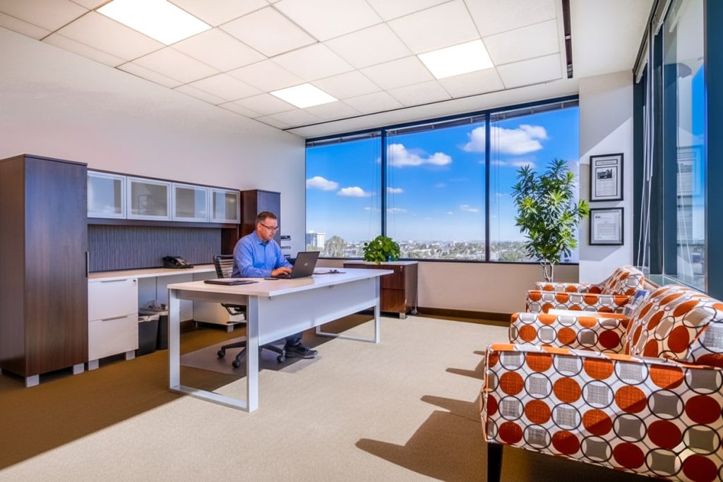 Barrister Suites Office Lease A man in a blue shirt works on a laptop at a modern office desk. Large windows show a city view under a blue sky. There are patterned chairs, plants, and office furniture in the bright, tidy room.
