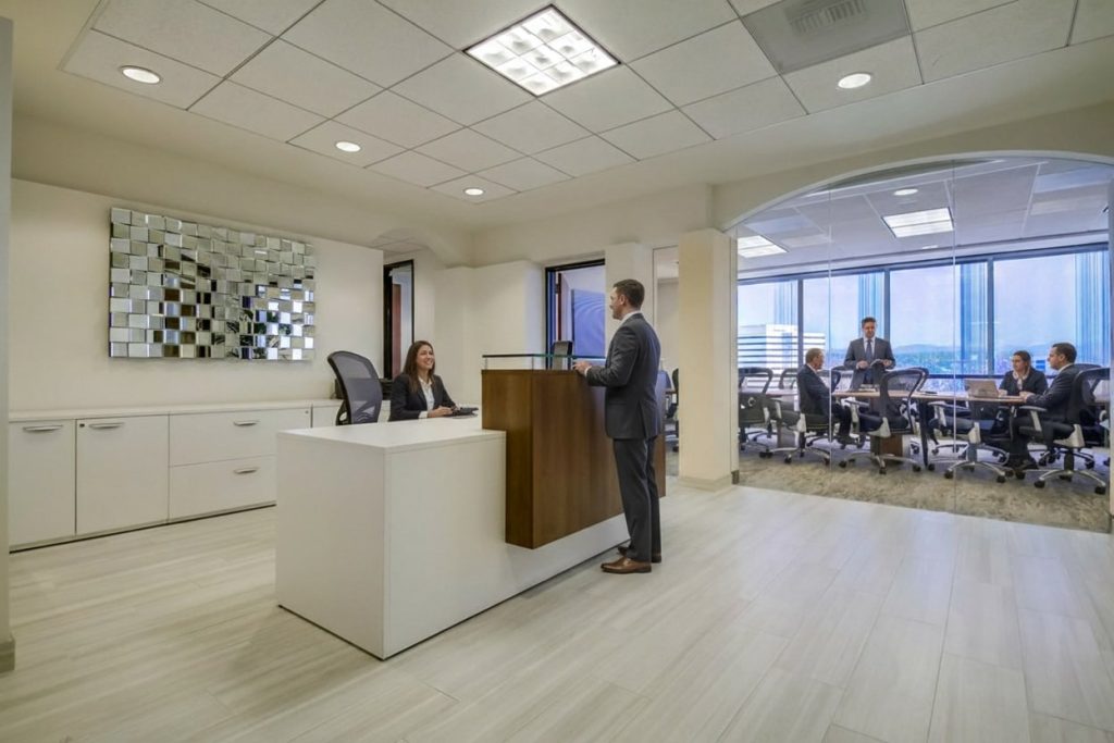 Barrister Suites Office Lease A modern office reception area with a woman seated at a desk speaking to a man in a suit. In the background, several people are gathered around a conference table near large windows with city views.