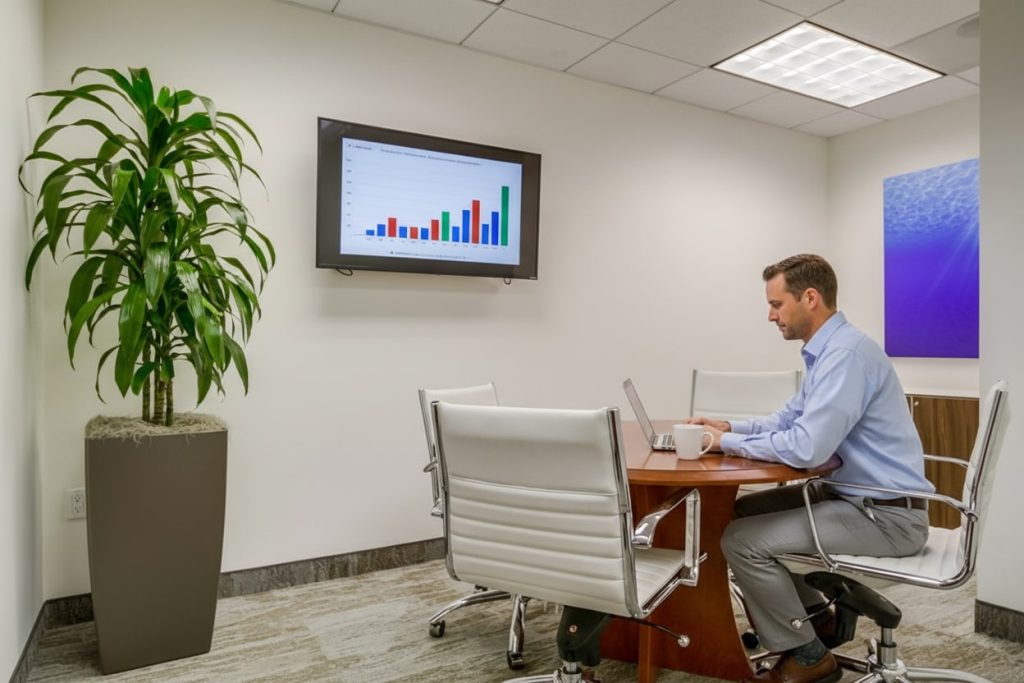 Barrister Suites Office Lease A man in business attire sits alone at a small conference table working on a laptop. A large plant is in the corner, and a bar graph is displayed on a wall-mounted screen. Four white chairs and a coffee mug are visible.