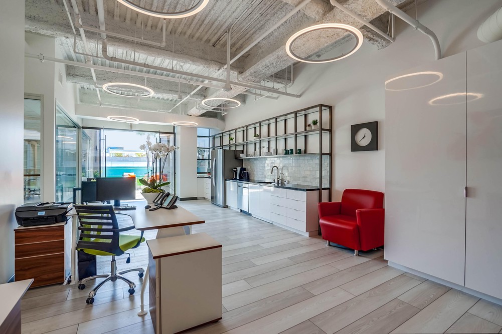 Barrister Suites Office Lease Modern office with light wood floors, white walls, circular ceiling lights, a red armchair, white cabinets, a desk with a green-accented chair, a printer, and shelves above a kitchenette. Large windows let in natural light.