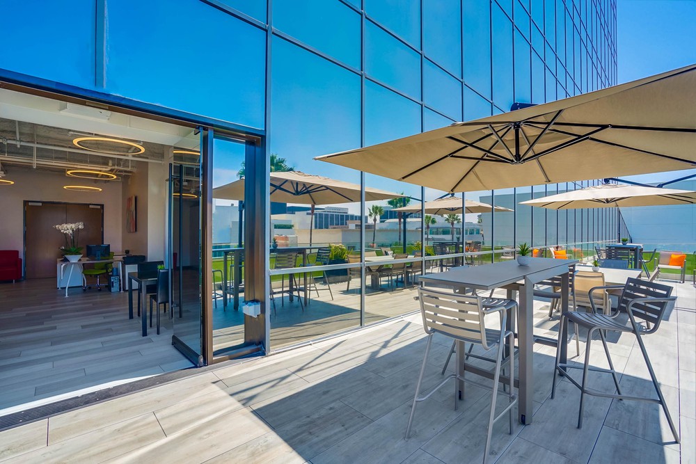 Barrister Suites Office Lease Modern outdoor patio with tall tables, chairs, and large umbrellas beside a glass-walled building. Indoor seating and greenery are visible through open sliding doors on a sunny day.