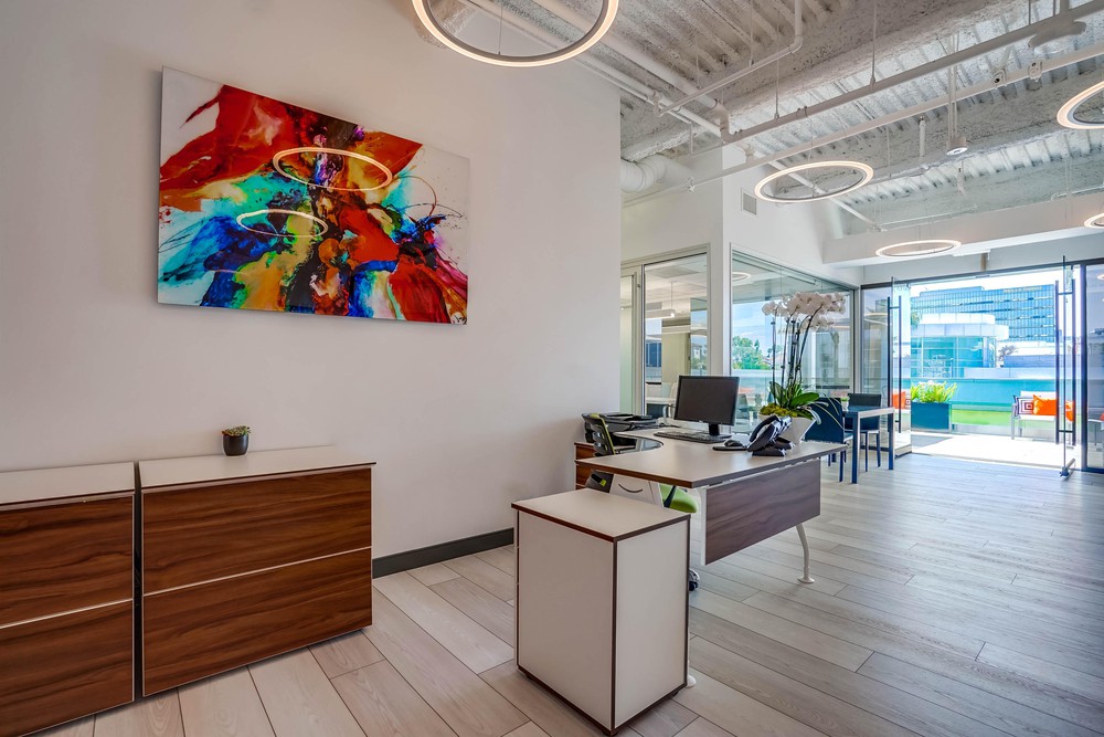 Barrister Suites Office Lease A modern office with a wooden desk, chairs, and cabinets. Colorful abstract art hangs on the wall. Circular ceiling lights and large windows let in natural light, with a view of a patio and cityscape outside.