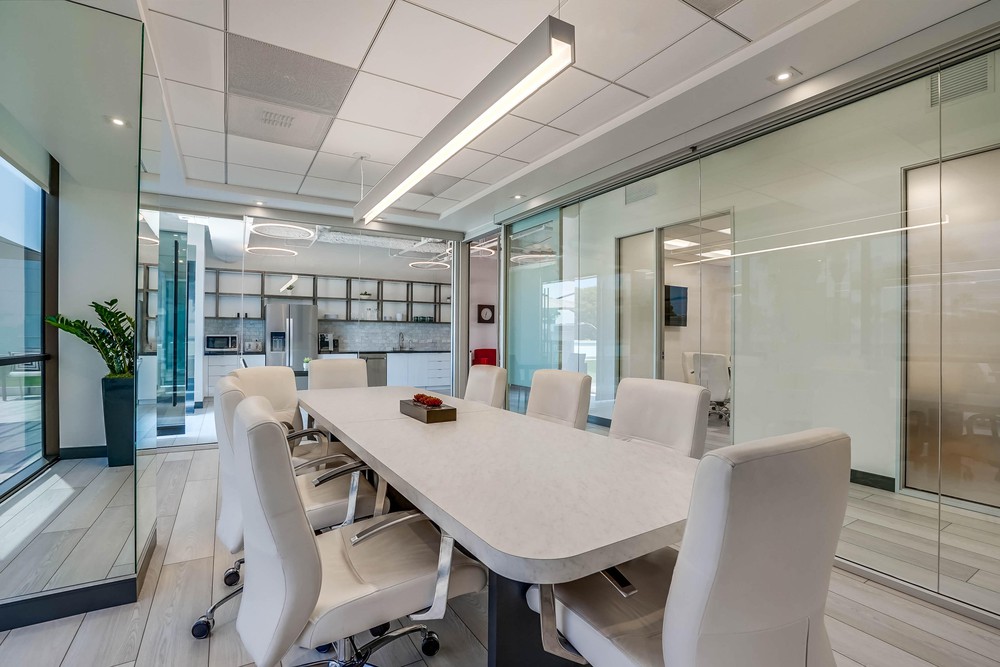 Barrister Suites Office Lease Modern conference room with a long white table, surrounded by eight white chairs, glass walls, and a view into a kitchen area. The space is bright, with contemporary lighting and a potted plant in the corner.