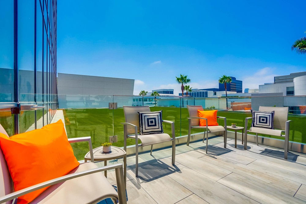 Barrister Suites Office Lease Modern outdoor patio with metal chairs featuring orange and black-and-white geometric pillows, glass railings, green lawn, and contemporary buildings in the background under a clear blue sky.