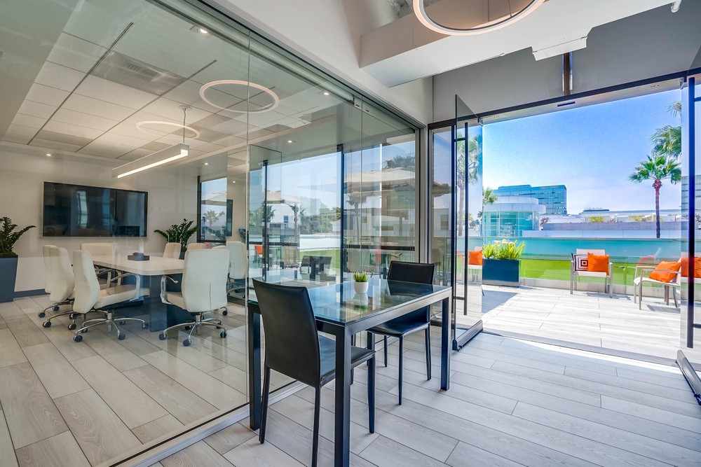 Barrister Suites Office Lease Modern office space with glass walls, a conference table and chairs inside, and a small table with black chairs near open glass doors leading to a balcony with bright outdoor seating and palm trees.