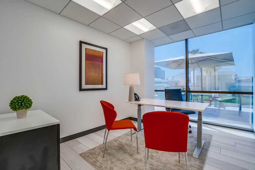 Barrister Suites Office Lease Modern office with a white desk, black office chair, and two red guest chairs. A lamp, framed artwork, and a small green plant decorate the space. Large window shows patio with umbrella and cityscape outside.