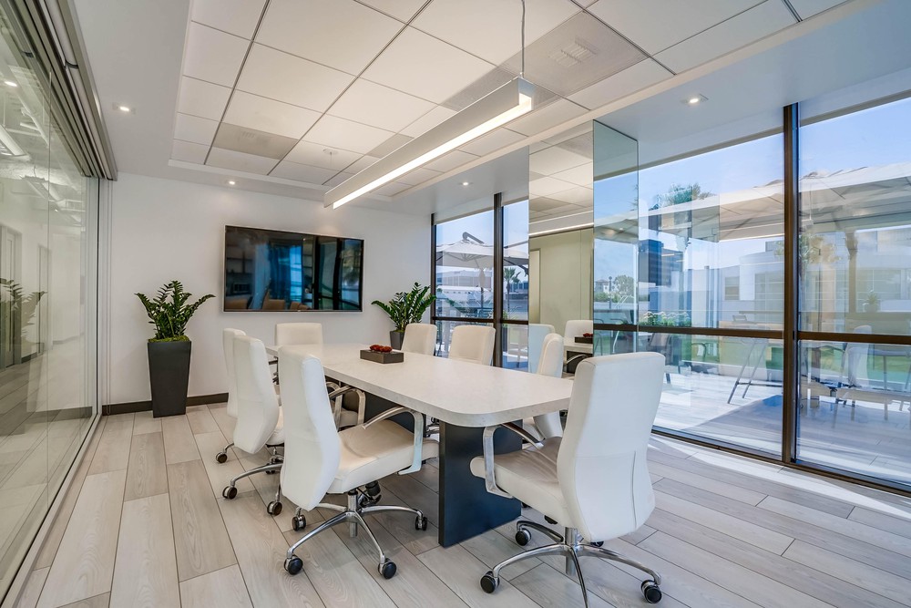 Barrister Suites Office Lease Modern conference room with a long white table, eight white chairs, a wall-mounted TV, two potted plants, glass walls, and large windows letting in natural light.