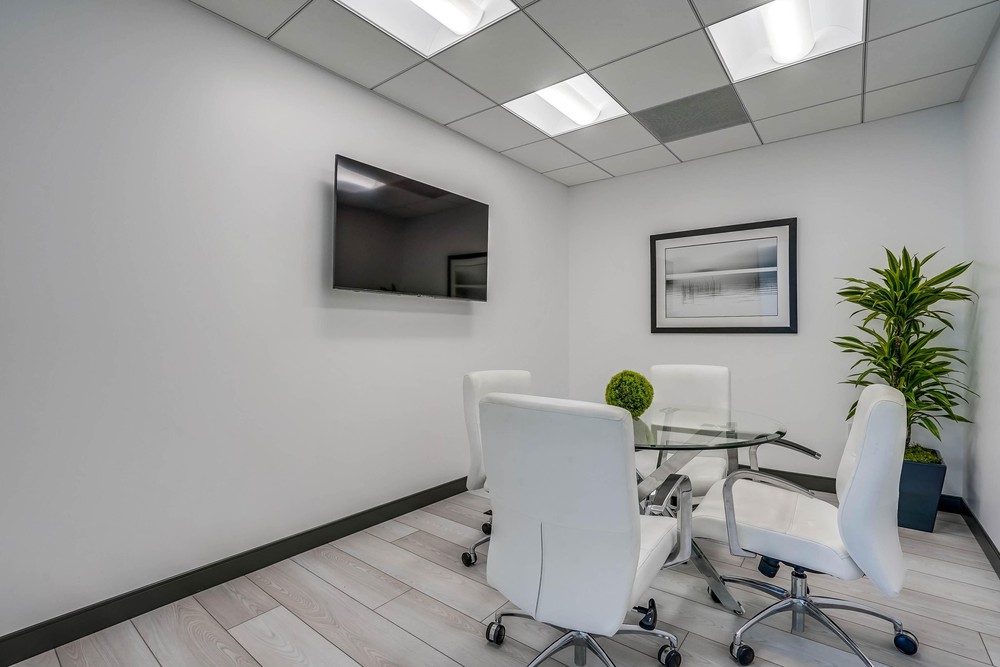 Barrister Suites Office Lease A small modern conference room with four white chairs around a glass table, a wall-mounted TV, a framed picture, a green plant in the corner, and light-colored flooring.