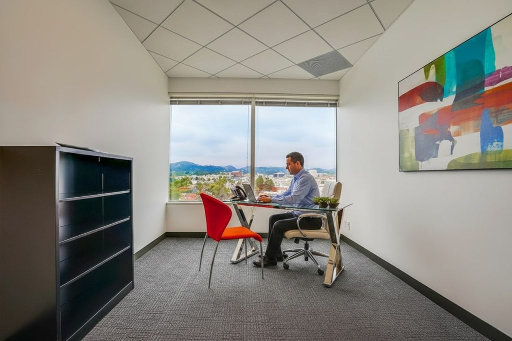 Barrister Suites Office Lease A man sits at a small desk in a modern office with a large window, working on a laptop. The room has minimal furniture, colorful abstract art on the wall, and a view of buildings and hills outside.