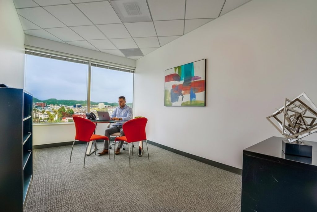 Barrister Suites Office Lease A man works on a laptop at a small desk with two red chairs in a modern office with large windows, abstract wall art, and geometric sculpture on a cabinet.