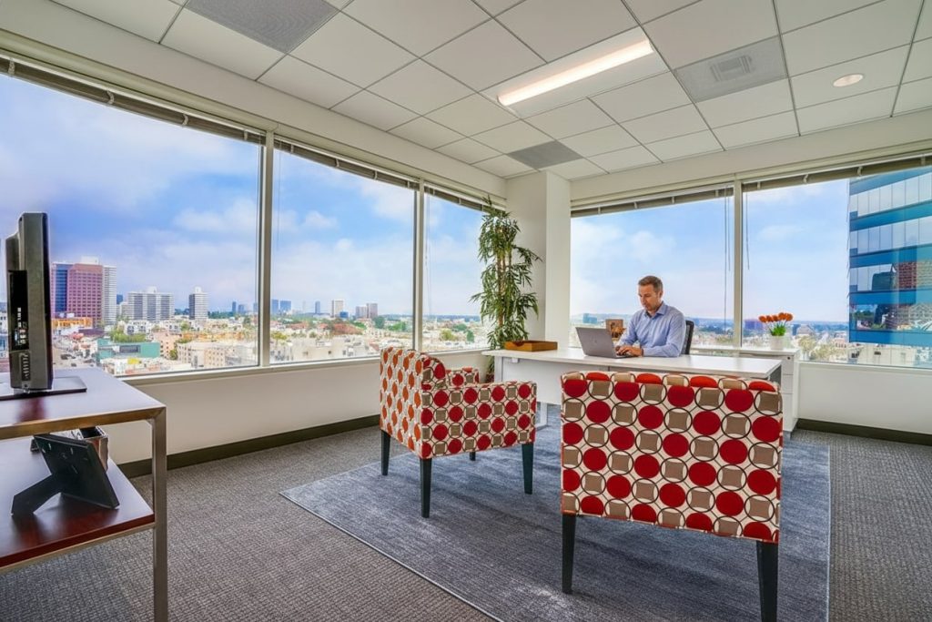 Barrister Suites Office Lease A man sits at a desk with a laptop in a modern office with large windows, patterned chairs, a plant, and a city skyline view in the background under a partly cloudy sky.