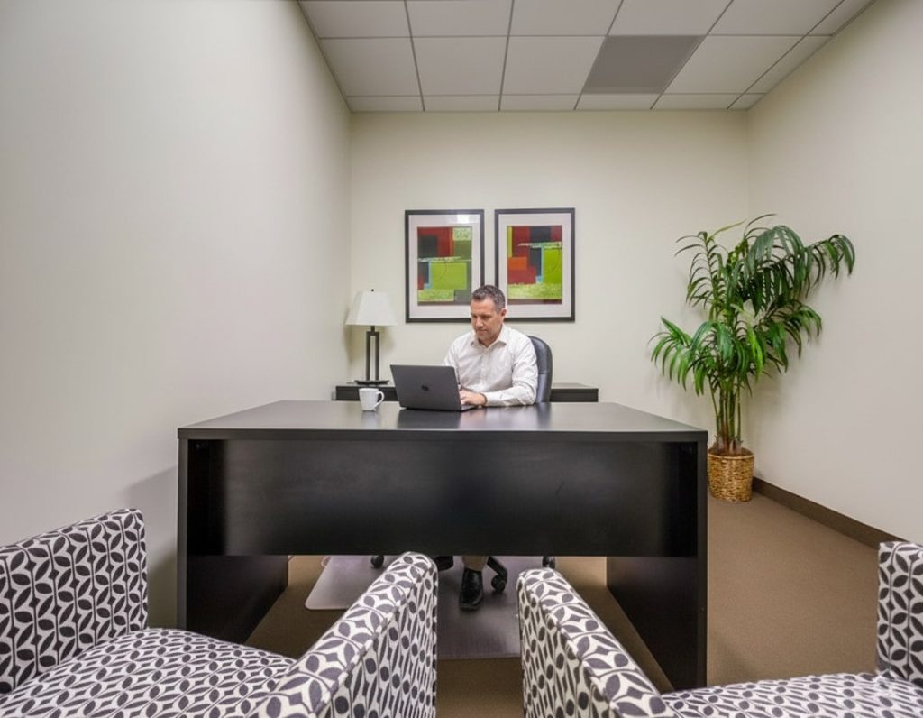 Barrister Suites Office Lease A man sits at a black desk working on a laptop in a small office with white walls, two patterned chairs, a potted plant, and two colorful abstract paintings on the wall.