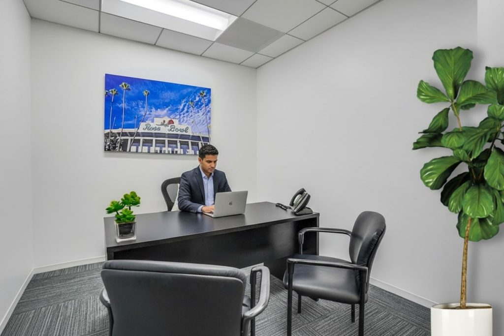 Barrister Suites Office Lease A man in a suit works on a laptop at a black desk in a modern, minimalist office with white walls, black chairs, two potted plants, and a colorful photo of the Santa Monica Bowl sign on the wall.