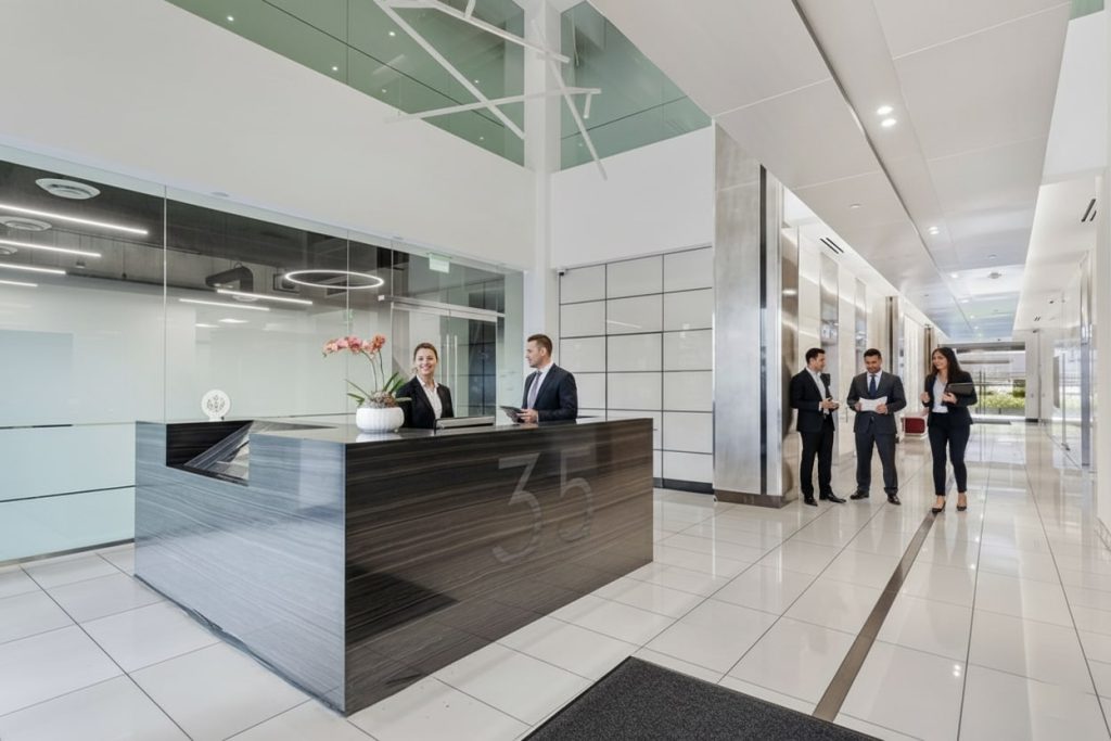 Barrister Suites Office Lease Modern office lobby with a dark reception desk where two receptionists are seated. Four people in business attire are having a conversation in the hallway. The space is bright with white floors and glass walls.