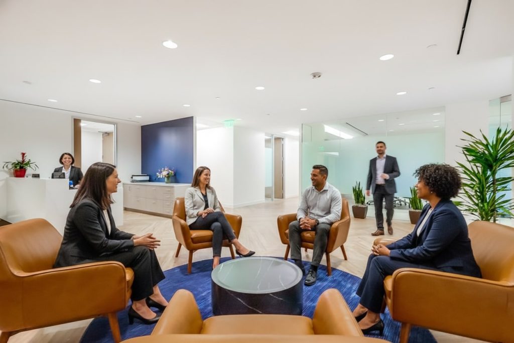 Barrister Suites Office Lease Five people in business attire are having a meeting, seated in a modern office lounge with brown chairs and a blue rug, while another person stands by a reception desk in the background.