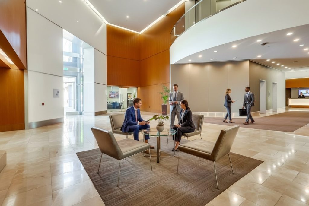 Barrister Suites Office Lease Business professionals in formal attire meet and talk in a modern office lobby with sleek furniture, marble floors, and high ceilings. Other people walk and work in the background, highlighting a corporate environment.