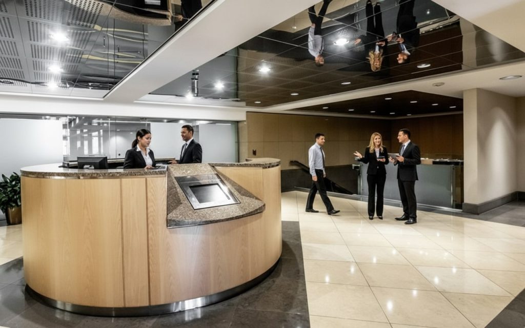 Barrister Suites Office Lease Spacious office lobby with a wooden reception desk; two people talk at the desk while three others stand and converse nearby. The ceiling reflects the scene below.