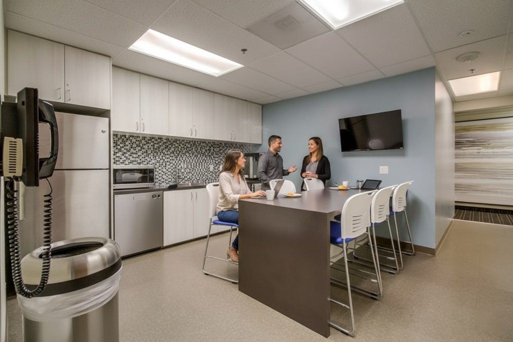 Barrister Suites Office Lease Three people talk in a modern office kitchen, with two seated at a counter and one standing. The room has white cabinets, a microwave, a fridge, and a wall-mounted TV. A trash can and phone are in the foreground.