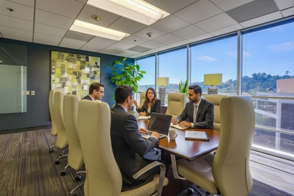 Barrister Suites Office Lease Four business professionals sit around a conference table in a modern office, discussing work. Large windows provide natural light and a scenic outdoor view, while office decor includes artwork and a potted plant.