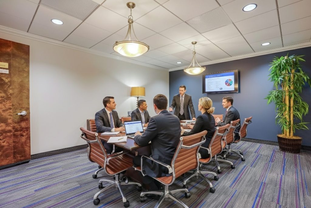 Barrister Suites Office Lease Six business professionals in suits sit around a conference table in a modern office, discussing documents and a laptop, while one stands by a screen displaying a colorful chart. A large plant is in the corner.