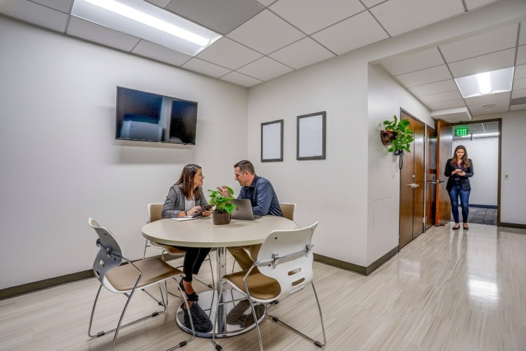 Barrister Suites Office Lease Two people sit at a round table in an office break area, talking and using a laptop. A plant and empty frames are on the wall. A woman stands in the hallway near an exit sign, holding a phone.