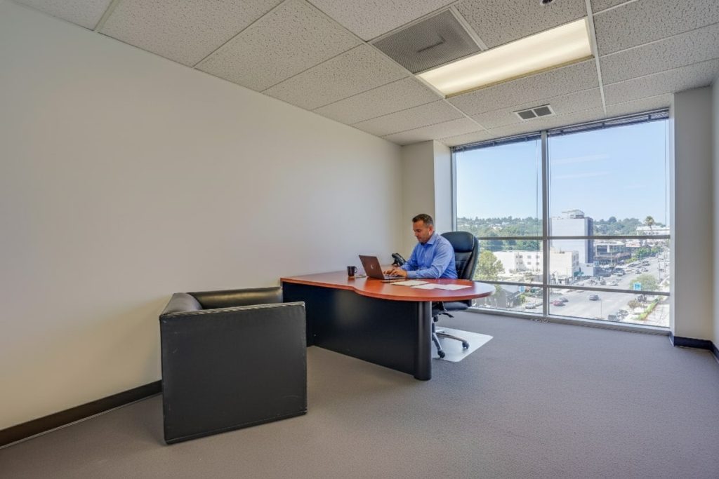 Barrister Suites Office Lease A man in a blue shirt sits alone at a desk in a spacious, modern office with large windows and a city view, working on a laptop. An empty black chair is in front of the desk.