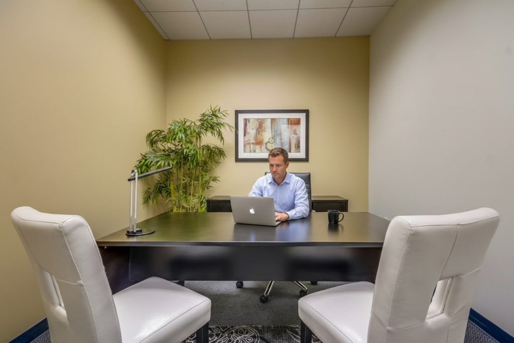 Barrister Suites Office Lease A man sits alone at a desk in a small office, working on a laptop. Two white chairs face his desk, a mug and desk lamp are on the table, and a potted plant and framed art hang on the beige walls.