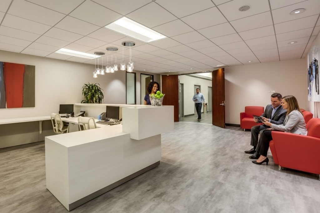 Barrister Suites Office Lease Modern office lobby with a white reception desk, a woman standing behind it, and three people sitting on red couches. Two men and a woman walk through a hallway in the background. The space is bright and tidy.