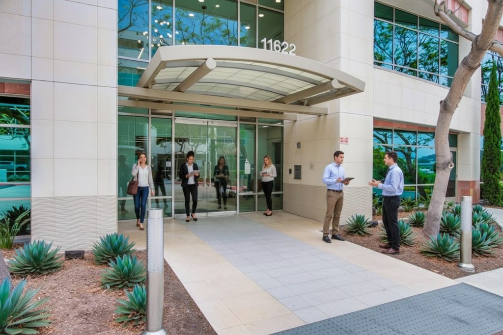 Barrister Suites Office Lease Several people, dressed in business attire, exit a modern office building with glass doors and an awning. Two men stand outside talking, while others walk along the pathway near landscaping with small plants.