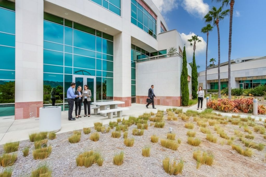 Barrister Suites Office Lease Four people stand and talk near a picnic table outside a modern office building with large windows, while another person walks by and a woman stands near plants holding a phone. Some landscaping with small shrubs is in the foreground.