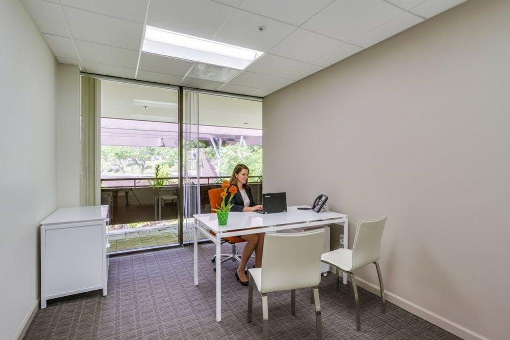Barrister Suites Office Lease A woman sits at a desk in a modern, minimalist office with two white chairs, a laptop, a phone, and a plant. Large windows behind her show a balcony and greenery outside.