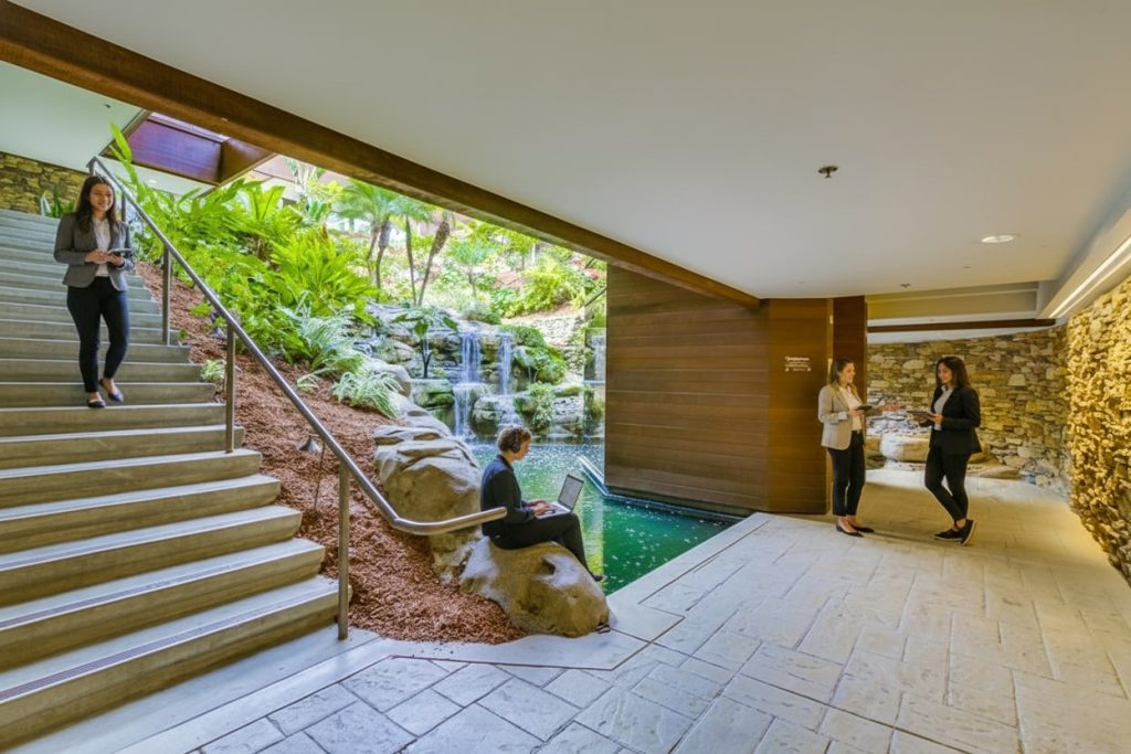 Barrister Suites Office Lease Four people in business attire are in a modern indoor space with a staircase, a small pond, rocks, and a lush green garden with a waterfall, blending natural and architectural elements.