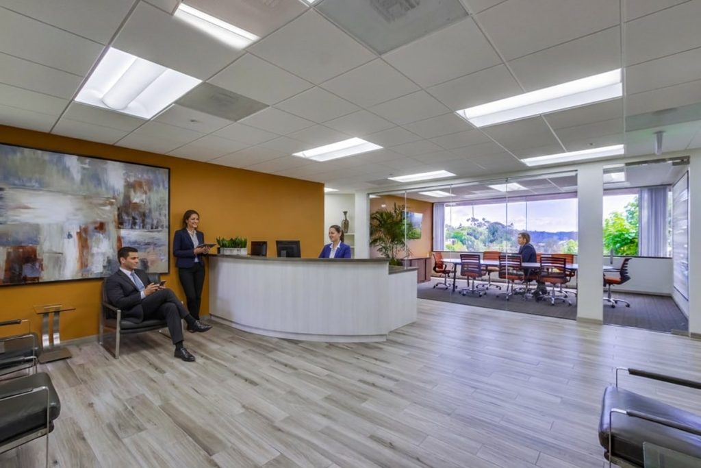 Barrister Suites Office Lease Modern office lobby with a curved reception desk, three people interacting, large abstract art on the wall, glass-walled conference room, and bright natural light from windows with a view of greenery outside.