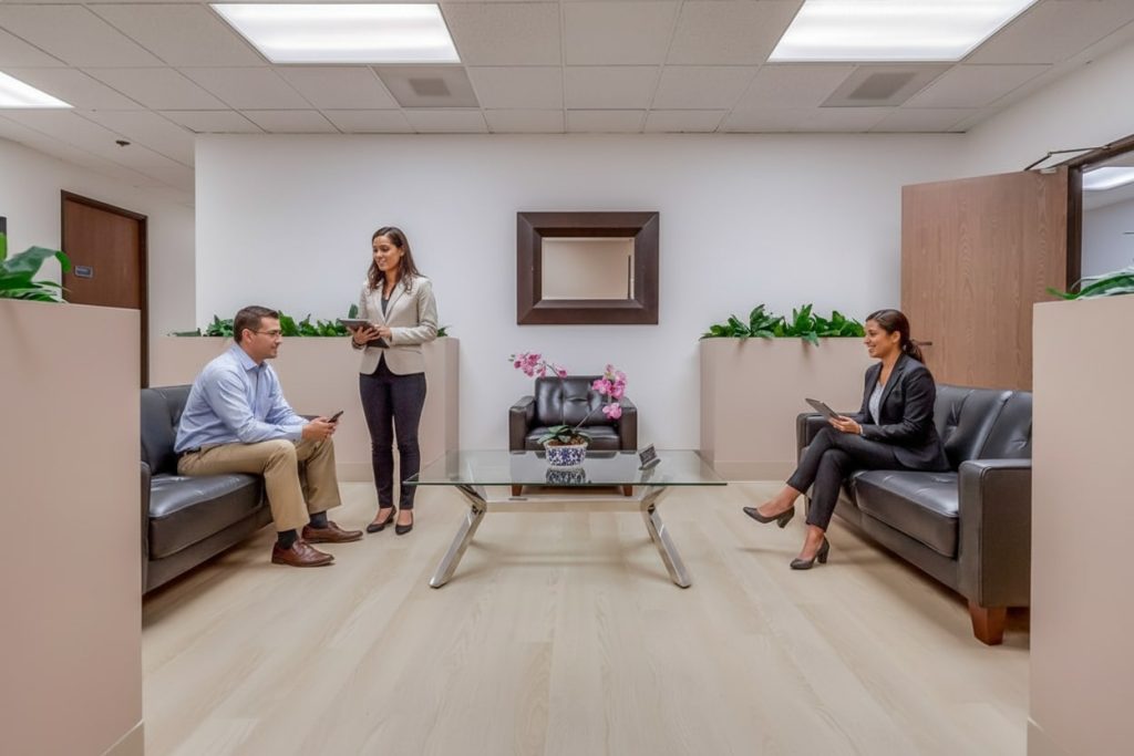 Barrister Suites Office Lease Three people in business attire are in a modern office waiting area with two black couches, a glass coffee table, a potted orchid, and wall plants. Two are seated with devices; one stands holding a clipboard.
