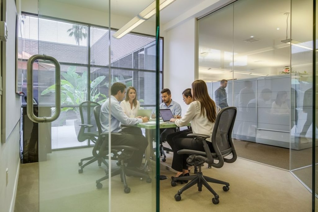 Barrister Suites Office Lease Five people in business attire sit around a table in a glass-walled office, engaged in a meeting. Laptops and notebooks are on the table. Bright, modern workspace with large windows and plants outside.