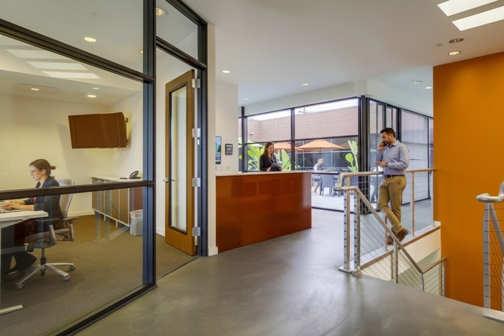 Barrister Suites Office Lease Modern office with glass-walled rooms; a woman works at a desk, another woman stands at a reception desk, and a man stands by a staircase talking on the phone. Large windows let in natural light.