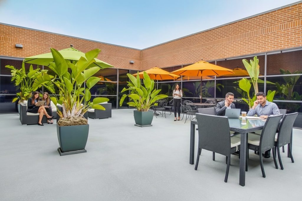 Barrister Suites Office Lease People sit and talk at outdoor tables with large green plants and orange umbrellas on a spacious patio next to a brick building. One woman stands and talks on her phone. The scene appears relaxed and modern.