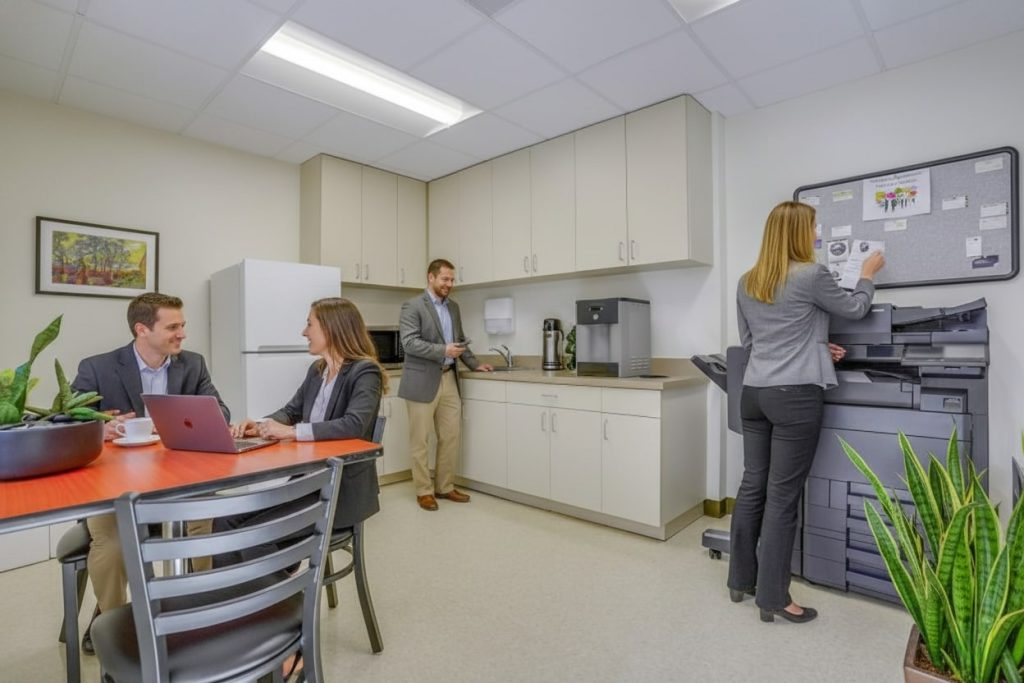 Barrister Suites Office Lease Four people in business attire are in an office break room. Two are sitting at a table with a laptop, one is getting coffee, and another is using a copier. The room has cabinets, plants, and a bulletin board.